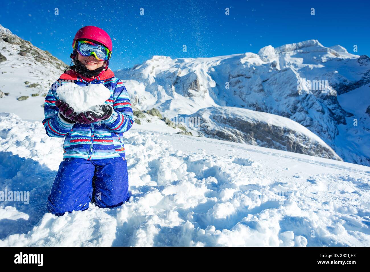 Sorridente ragazza carina felice in maschera da sci e casco mostra amore concetto di tenuta neve forma in cuore in piedi sulle ginocchia con montagne sulla parte posteriore Foto Stock