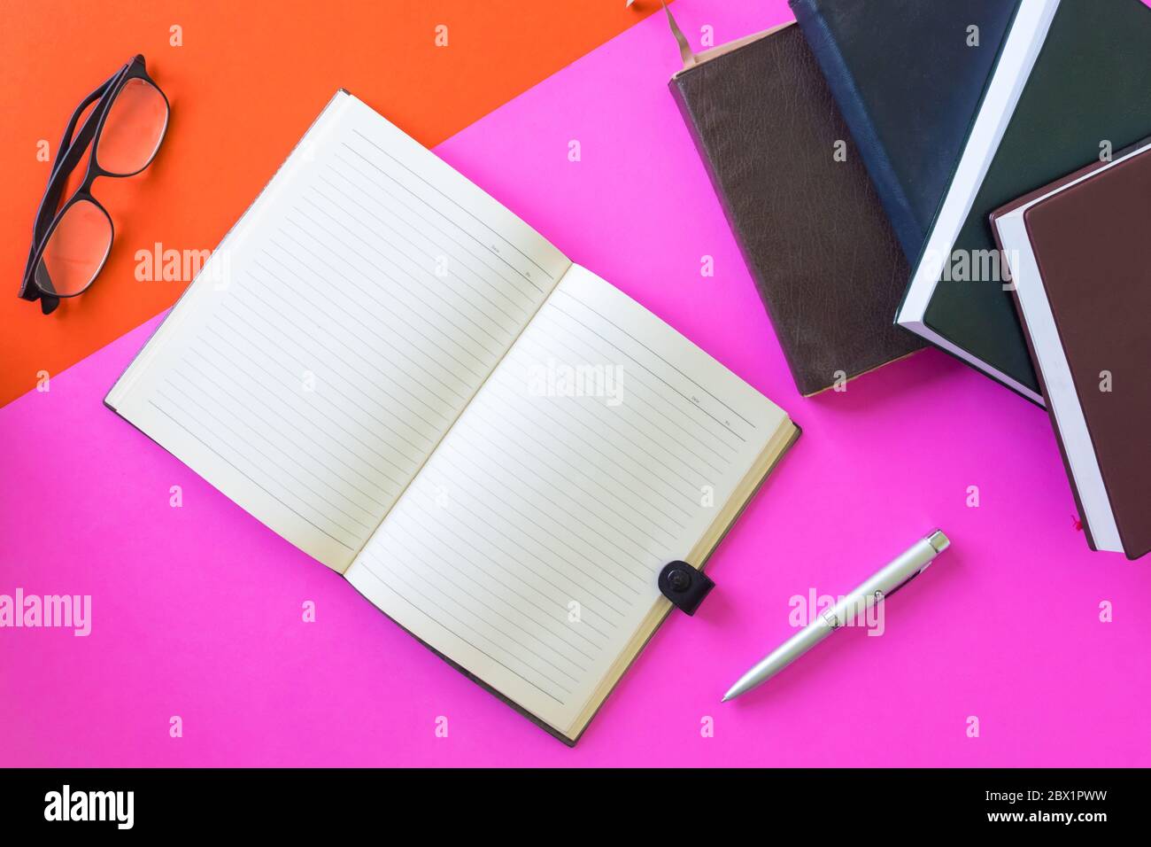 Apri un notebook vuoto con penne, occhiali e libri hardback su sfondo colorato. Foto Stock