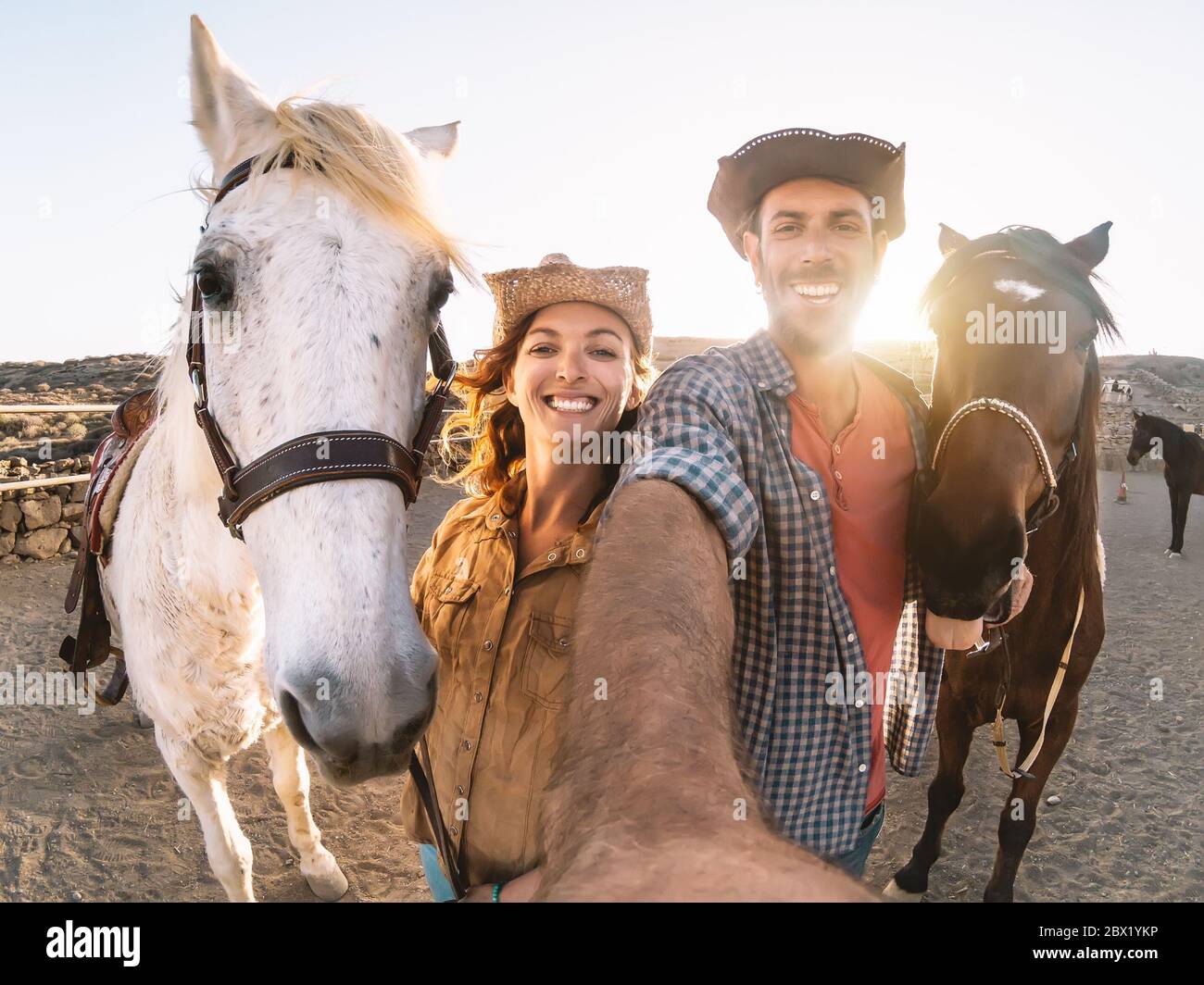 Felice coppia che prende selfie con cavalli all'interno della scuderia - giovani agricoltori che si divertono con gli animali nel ranch corrale Foto Stock