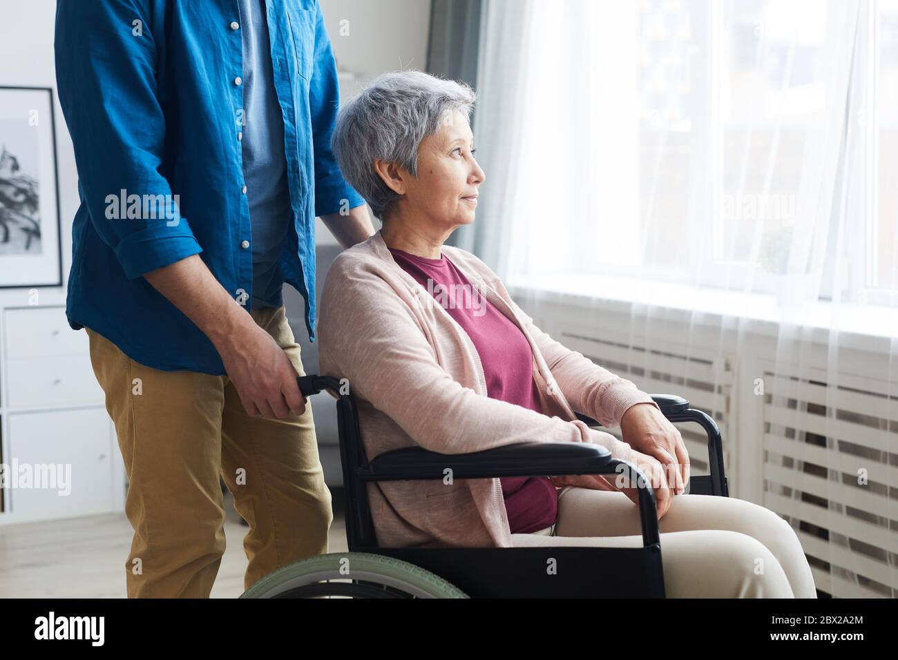 Donna anziana con disabilità seduta in sedia a rotelle e guardando attraverso la finestra con operatore sanitario in piedi nelle vicinanze Foto Stock