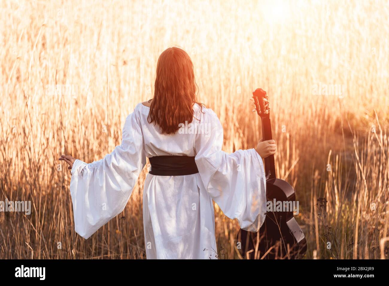 Vista posteriore di giovane donna snella con capelli rossi in un vestito bianco medievale e cintura che tiene la chitarra acustica e che cammina attraverso il campo di luce del tramonto. Foto Stock