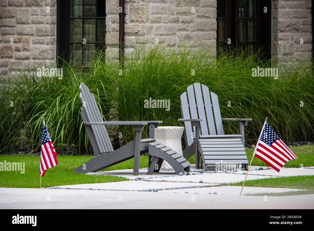 Due sedie grigie Adirondack seduta su un piccolo patio con tavolo tra loro di fronte a erba decorativa alta e finestre di casa di roccia - Fla americana Foto Stock