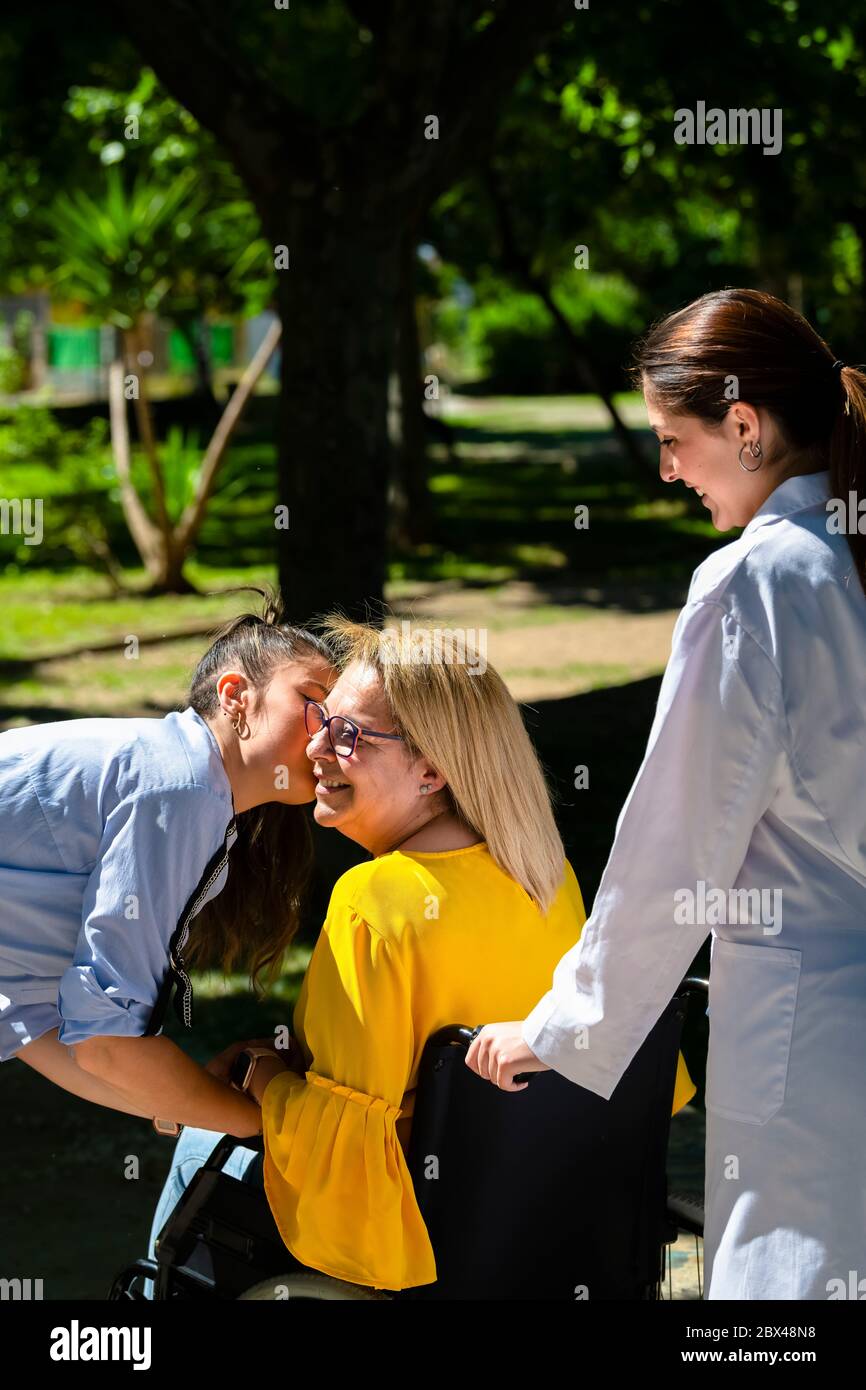 Una donna matura in sedia a rotelle sorride mentre viene baciata sulla guancia da un'altra donna su uno sfondo fuori fuoco. Concetto di salute e amicizia. Foto Stock