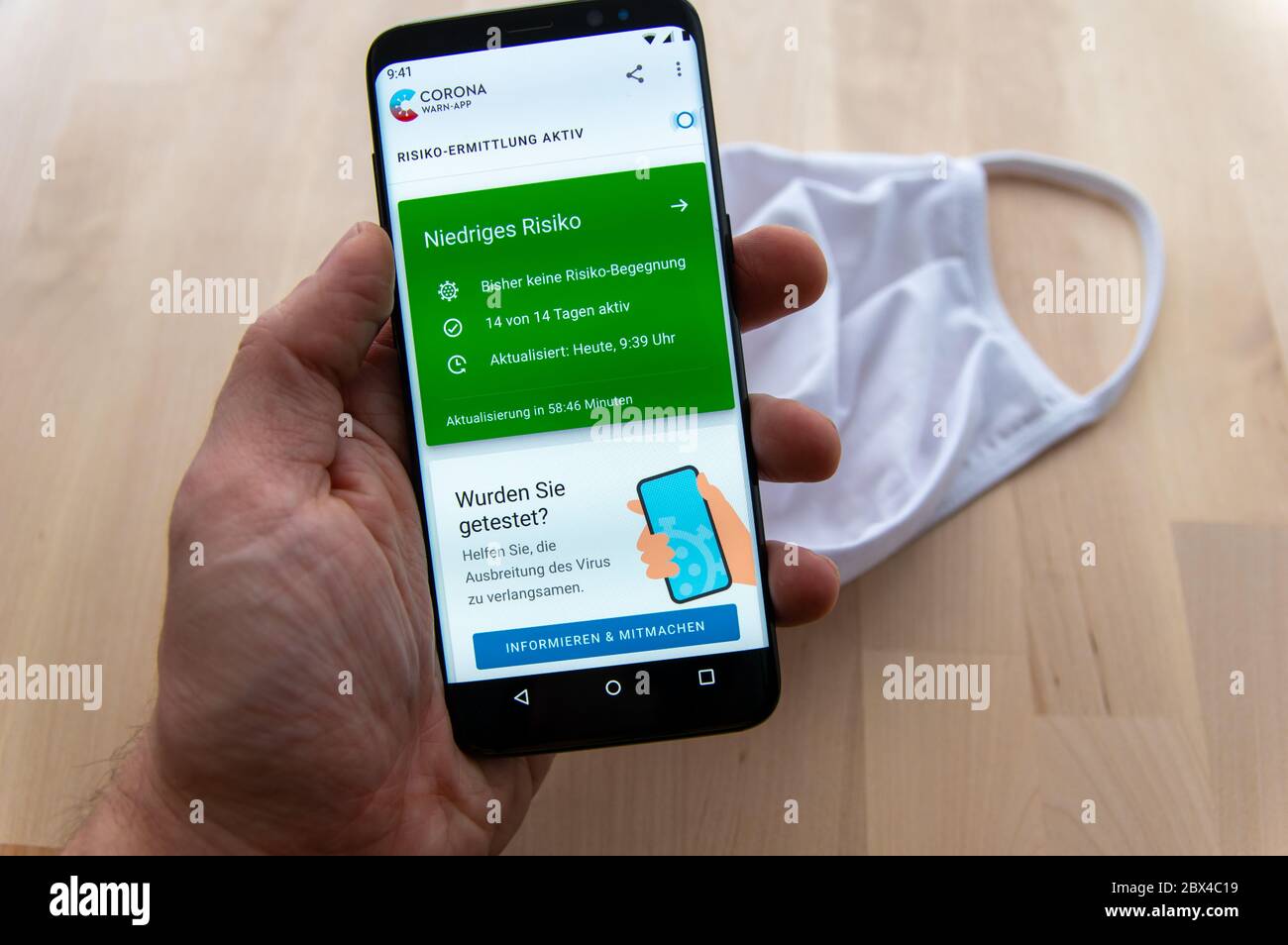 Zittau, Sassonia / Germania - 5 giugno 2020: Smartphone con tenuta a mano che mostra l'analisi attiva del rischio dell'applicazione tedesca Corona WARN con maschera bianca sfocata Foto Stock