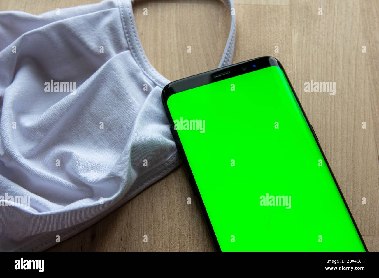 Display verde vuoto di uno smartphone accanto a una maschera bianca sulla superficie di legno. Modello ideale per Corona Tracing App Mockup. Foto Stock