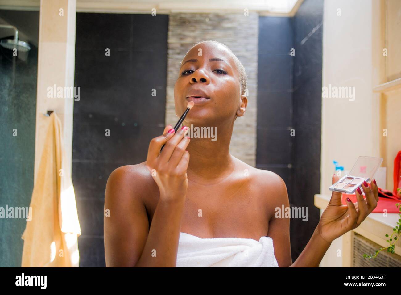 Stile di vita ritratto naturale di giovane attraente e felice donna afroamericana nera a casa bagno applicando trucco viso con pennello guardando fatica Foto Stock