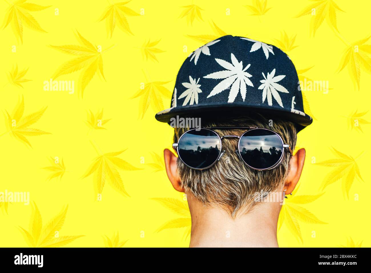 Ragazza in occhiali da sole, baseball con foglie marijuana fuma su sfondo giallo Foto Stock