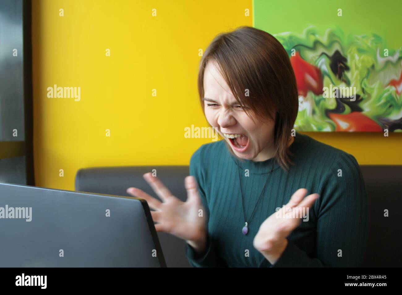 Una ragazza con un'espressione furiosa di tensione sul suo volto si siede lavorando a un computer portatile a un tavolo in un bar su un divano marrone contro una parete gialla. Guarda lo schermo del computer, urla e fa ondate le mani. Foto Stock