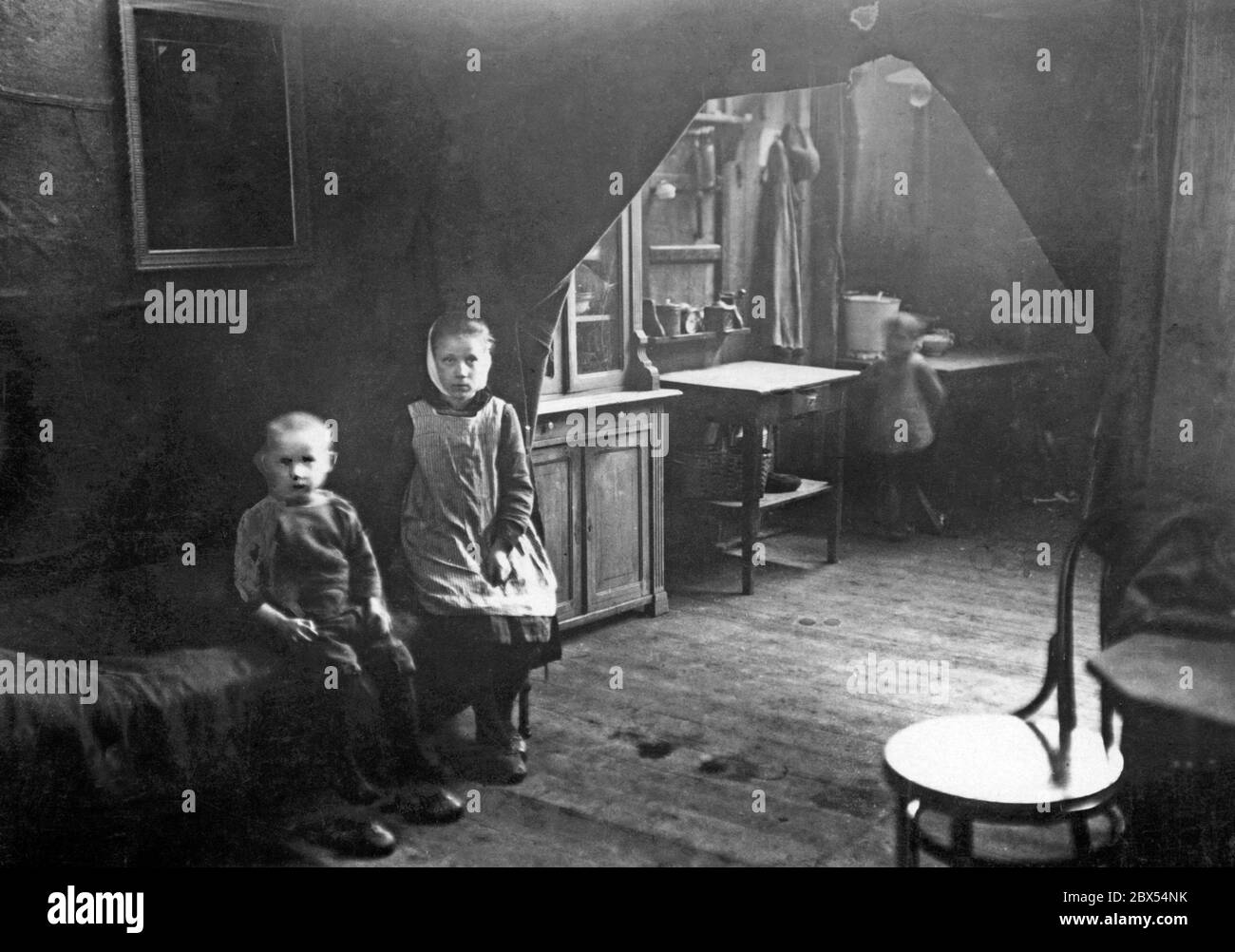 Due bambini si siedono su un letto in una casa buia. Alla loro destra è la cucina in cui un altro bambino (offuscato) sta in piedi. La cucina e la zona notte sono separate da tende. A causa dell'occupazione francese, nella regione del Reno-Ruhr vi era una carenza di alloggi. Foto Stock