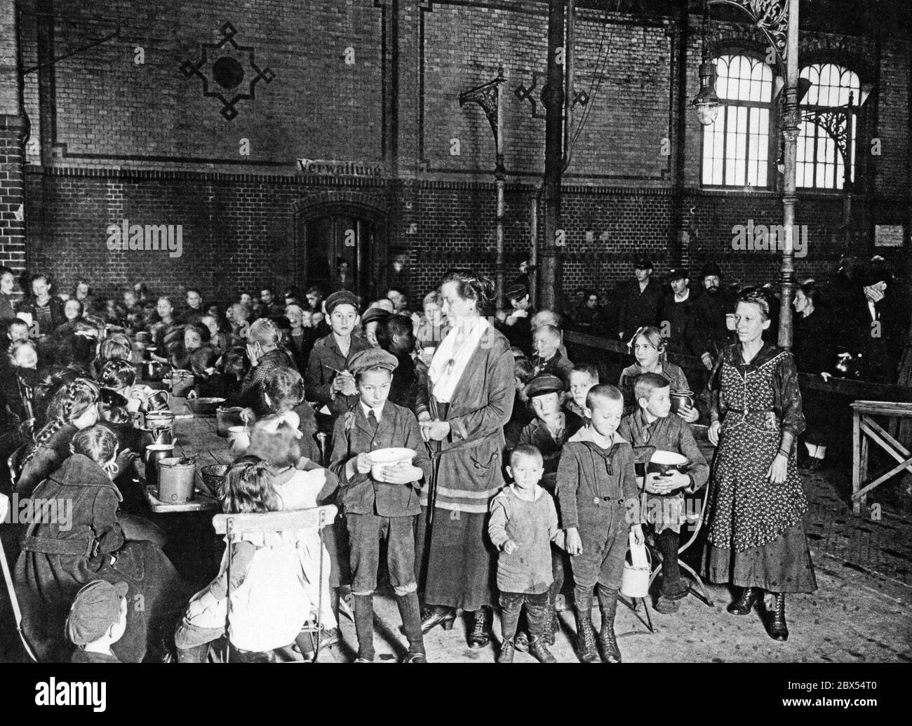 Gli studenti di una scuola elementare di Berlino siedono a tavoli lunghi in una grande sala. Davanti a loro sono grandi pentole di cibo. Due donne, presumibilmente le insegnanti, controllano i bambini. I pasti scolastici giornalieri sono sostenuti da donazioni straniere. Foto Stock