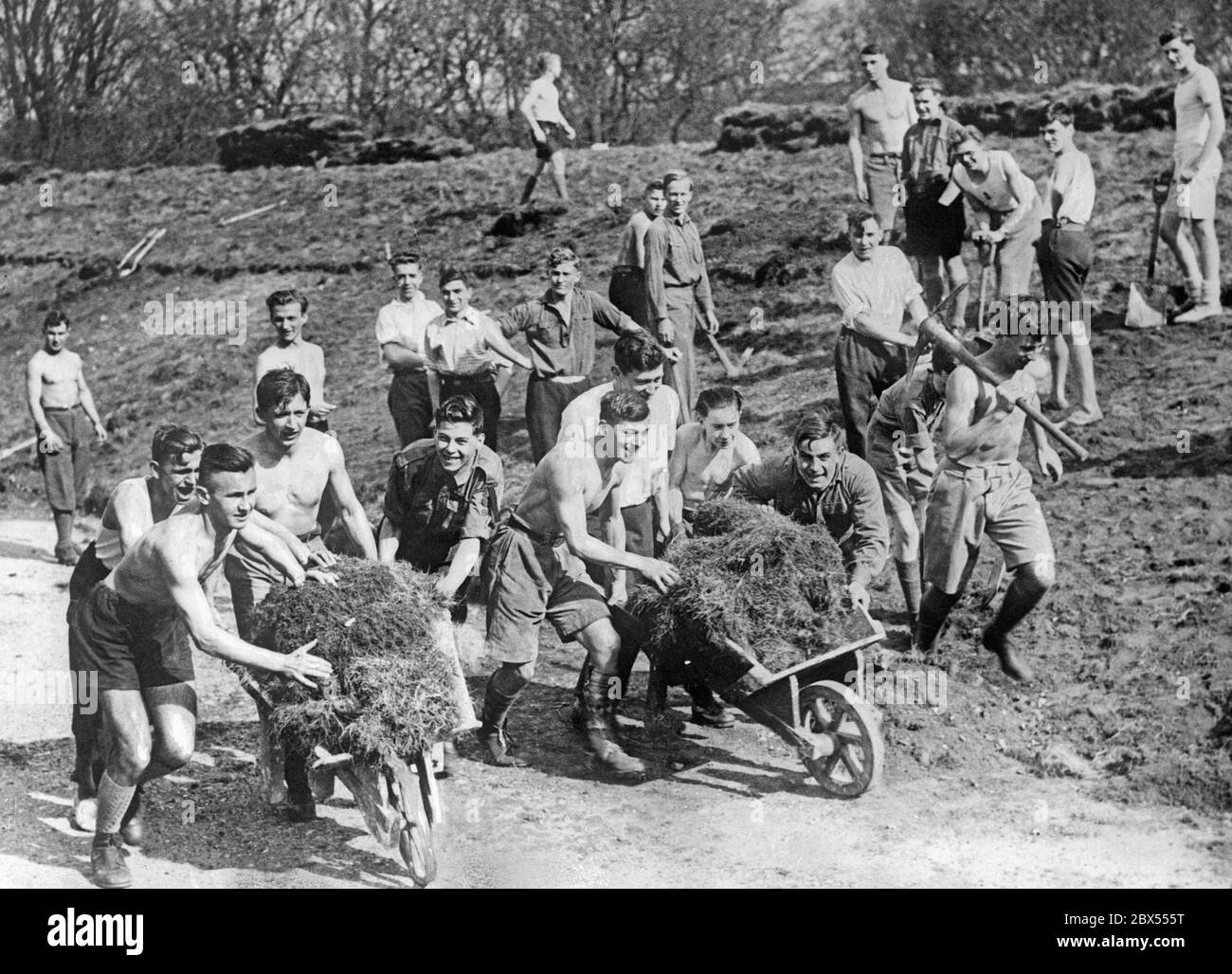 Ragazzi inglesi e tedeschi spingono i carrieri con fieno insieme in un campo comunale a Blandford. Alcuni trasportano pickasse e pale. Foto Stock