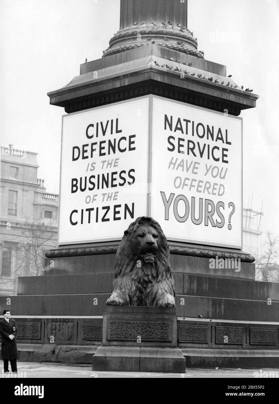 I poster pubblicitari 'Difesa civile' e 'Servizio Nazionale' sono collocati alla base della colonna Nelson in Trafalgar Square. In primo piano, una delle statue leone. Foto Stock