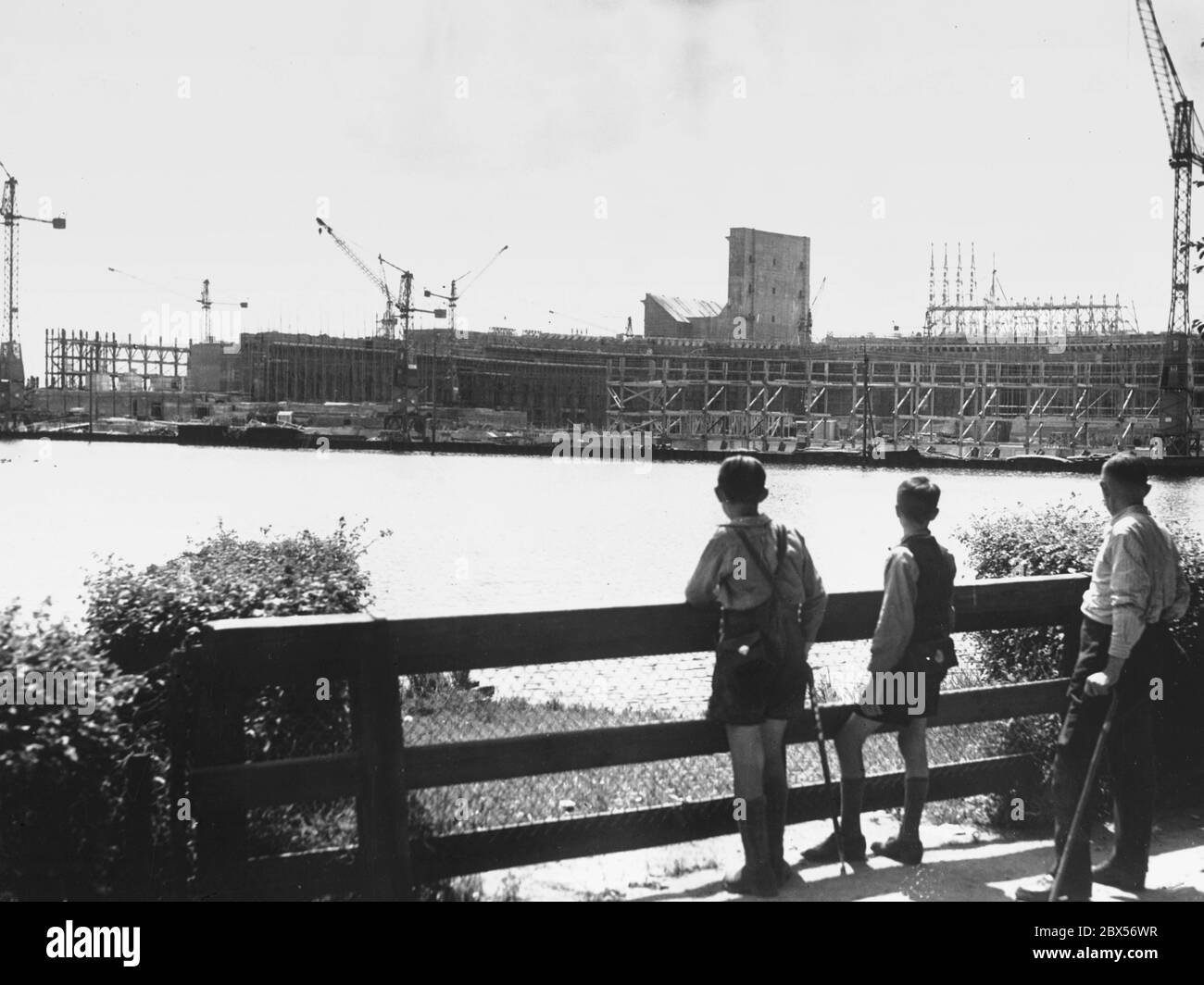 Un uomo (con la stampella) e due ragazzi guardano attraverso la Grosse Dutzendteich al cantiere della Sala Congressi progettato dagli architetti Ludwig Ruff e Franz Ruff sul terreno del Rally del Partito nazista a Norimberga. Foto Stock