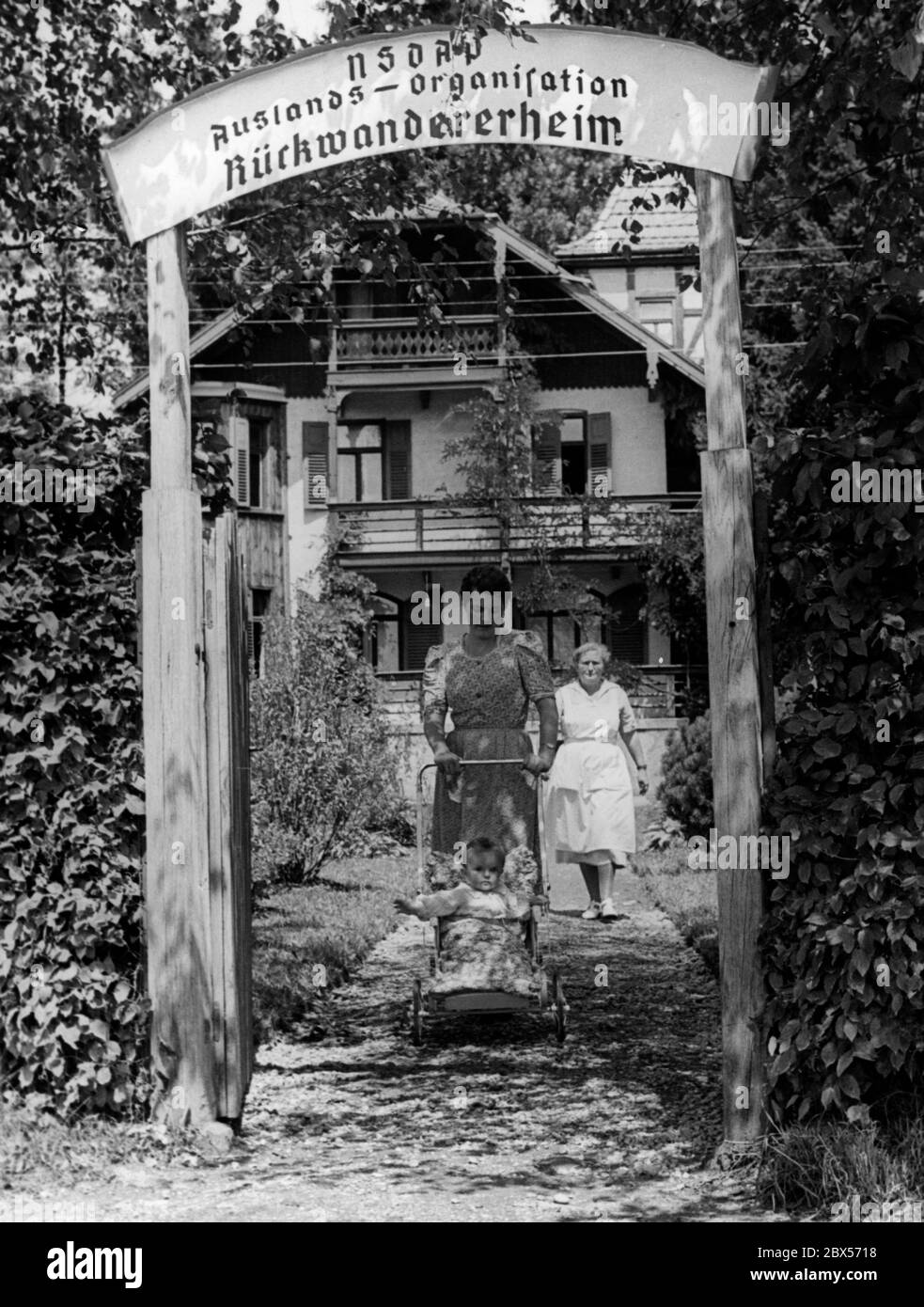 Una madre lascia con il suo bambino la Casa dei Bambini dell'Organizzazione degli Esteri della NSDAP, per fare una passeggiata nella vicina foresta. Dietro, davanti alla casa per i rimpatriati, c'è una donna in un cappotto bianco. Foto Stock