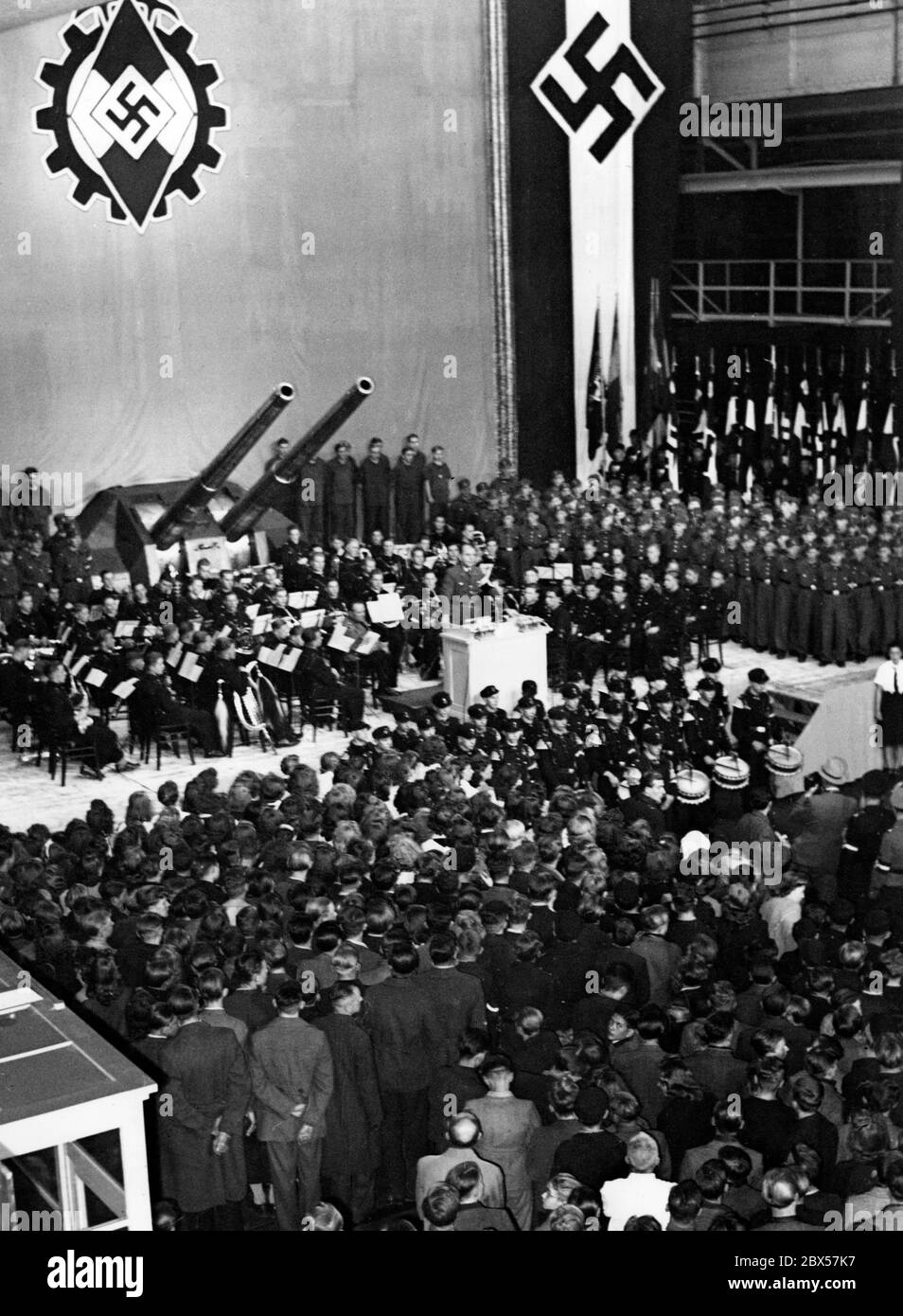 La forza lavoro in diminuzione dovuta alla "chiamata del fronte" è completata da apprendisti e giovani, nonché da lavoratori forzati e volontari dall'estero. Il Ministro degli armamenti Albert Speer doveva mantenere in vita l'economia bellica della guerra totale e quindi pretendeva "uno sforzo di lavoro incondizionato". La swastika centrale dietro le due pistole rappresenta una sovrapposizione delle bandiere del fronte di lavoro tedesco (DAF) e della gioventù Hitler (HJ). Foto Stock