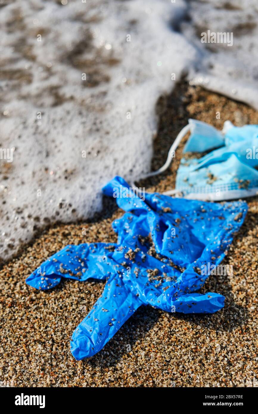 primo piano di una maschera chirurgica blu usata e un guanto di lattice blu gettato sulla sabbia bagnata della riva di una spiaggia Foto Stock
