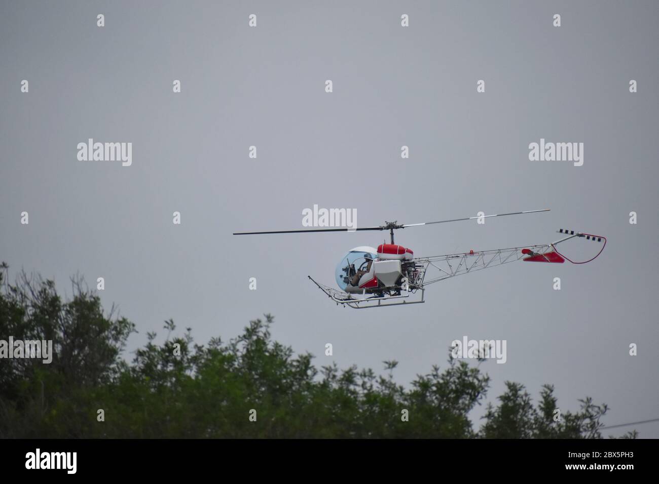 Elicottero a volo basso che si trova appena sopra gli alberi in una giornata nuvolosa con la silhouette di pilota in casco Foto Stock