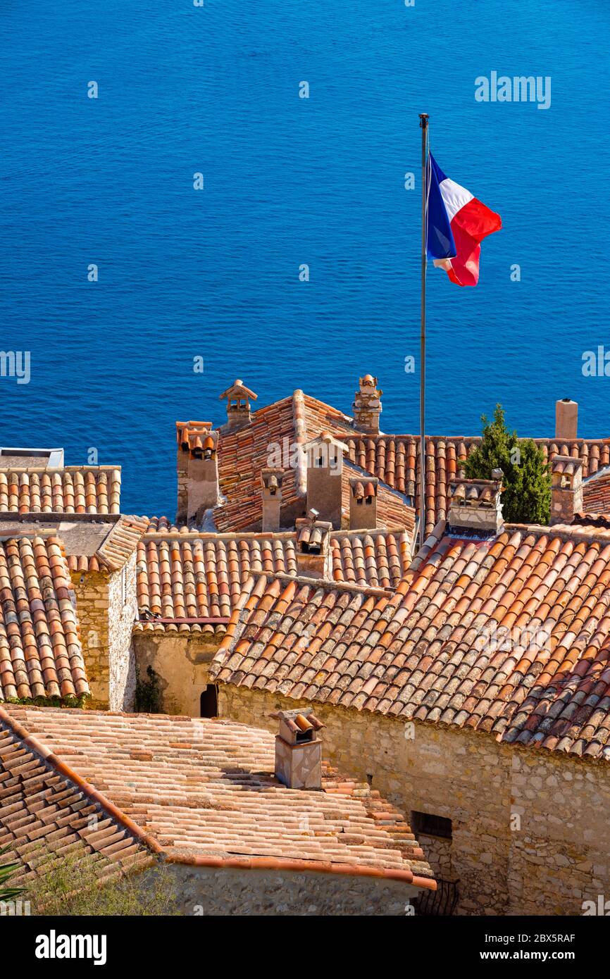I tetti in tegole di terracotta del villaggio di Eze con bandiera francese e il Mar Mediterraneo. Costa Azzurra, Alpi Marittime (06), Francia Foto Stock
