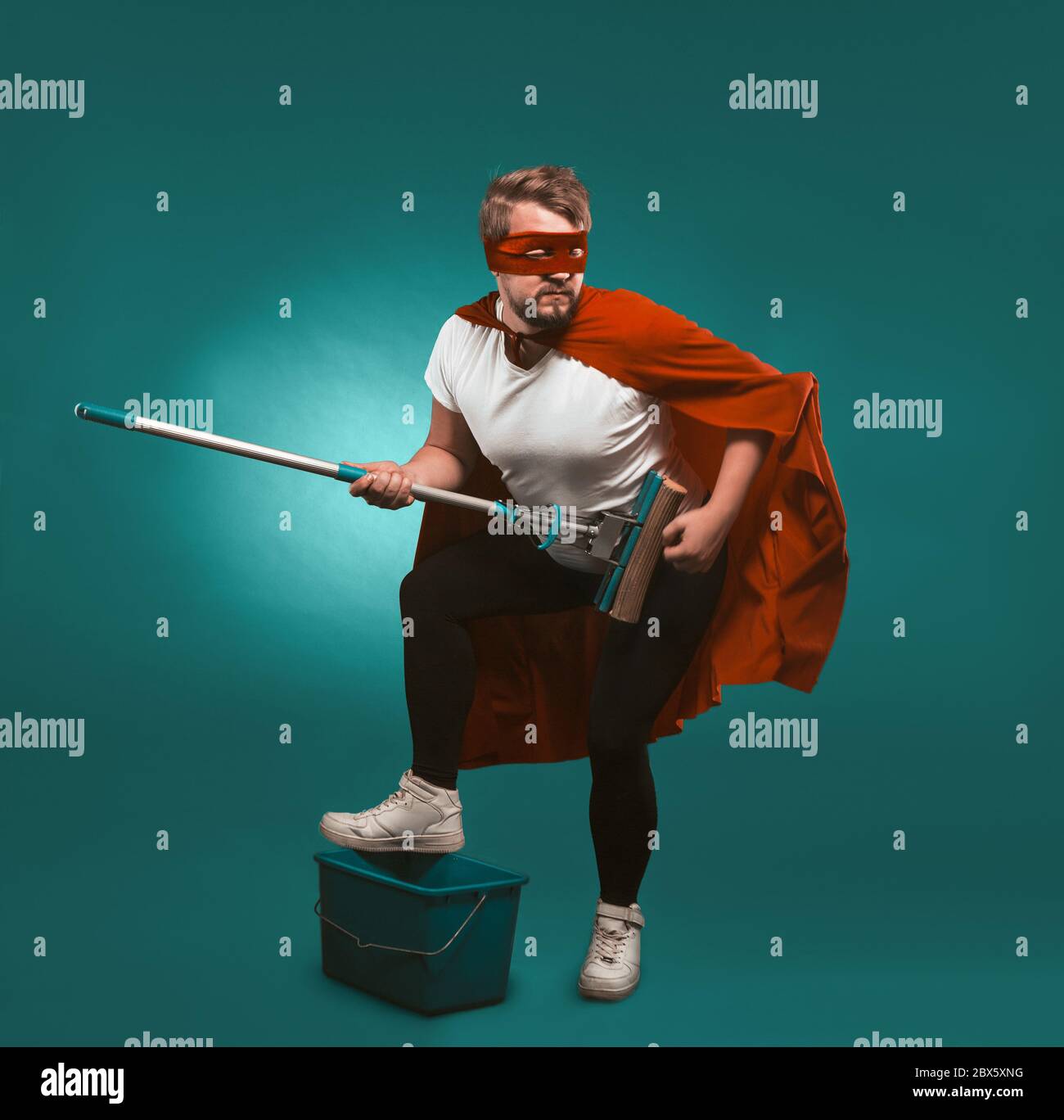 Il musicista rock supereroe sta pulendo. L'uomo che indossa un mantello rosso e una maschera tiene una mop che si rappresenta un musicista che mette il piede su un secchio. Taglia Foto Stock