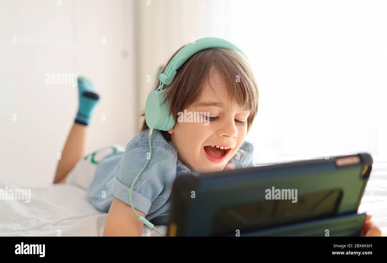 Il bambino moderno in cuffia usa un tablet digitale e sorride mentre si sdraia sul letto a casa: Carina un bambino in una camicia blu giocando a giochi e acquazzone Foto Stock