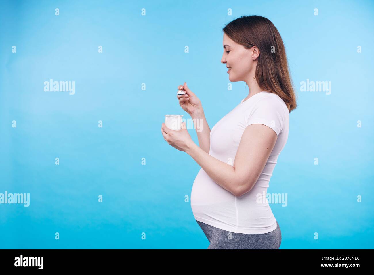 Vista profilo di giovane donna in gravidanza sana in camicia bianca mangiare yogurt da piccola tazza di plastica mentre si trova su sfondo blu Foto Stock