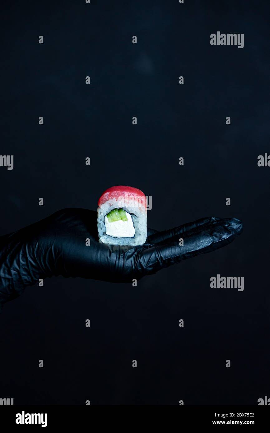 Mano femminile in guanti di gomma che tengono un rotolo di tonno sushi su uno sfondo scuro Foto Stock