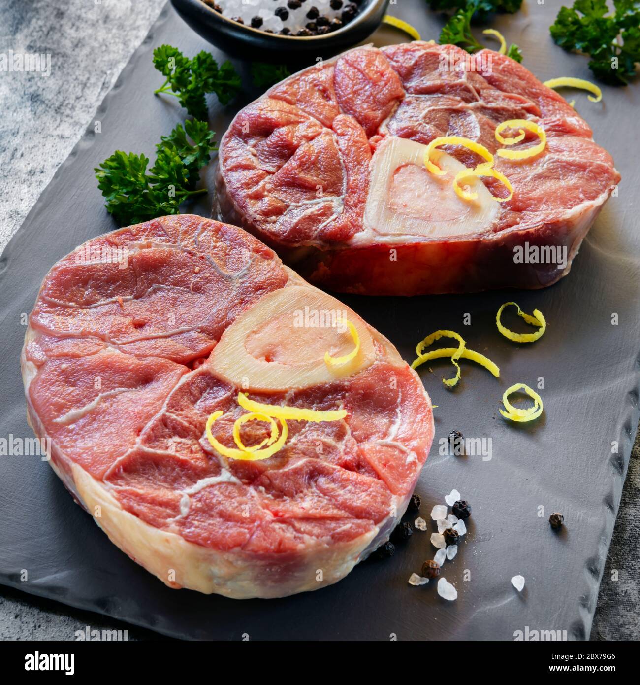 Gambi di vitello di osso buco, crudi, su ardesia. Con sale, pepe, prezzemolo e scorza di limone per la gremolata. Foto Stock