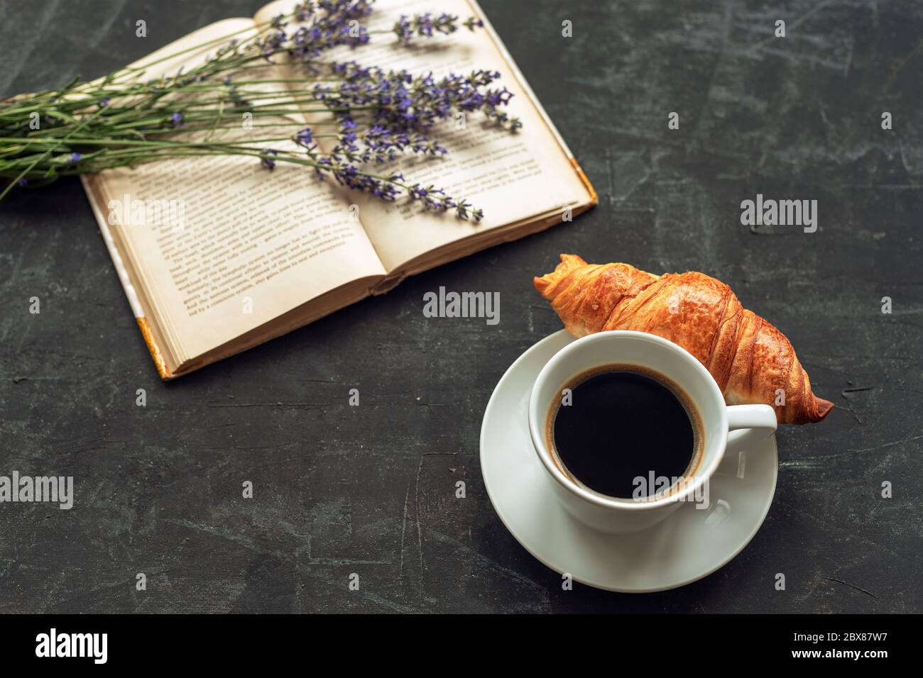 Tazza di caffè con croissant, libro e bouquet di lavanda su tavola nera. Foto Stock