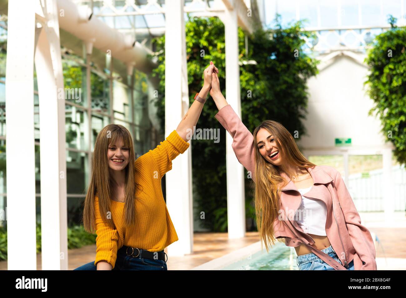 Due ragazze caucasiche giovani attraenti con capelli biondi scuotono le mani con le braccia in aria sorridendo ad una fontana all'interno di una stanza luminosa con il Foto Stock