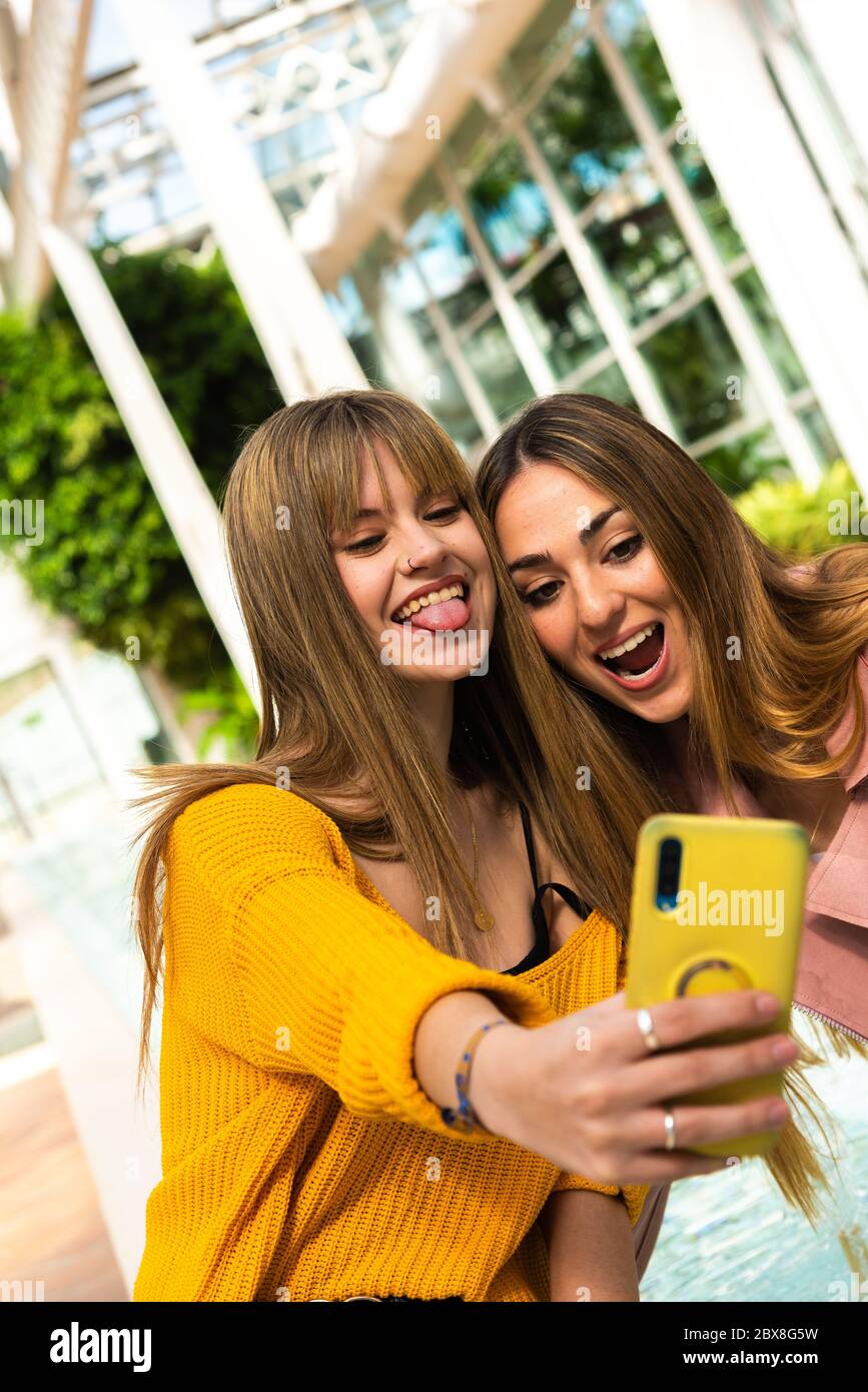 Due giovani ragazze caucasiche attraenti con i capelli biondi sono fotografate con il loro telefono cellulare in una fontana all'interno di un edificio bianco luminoso Foto Stock