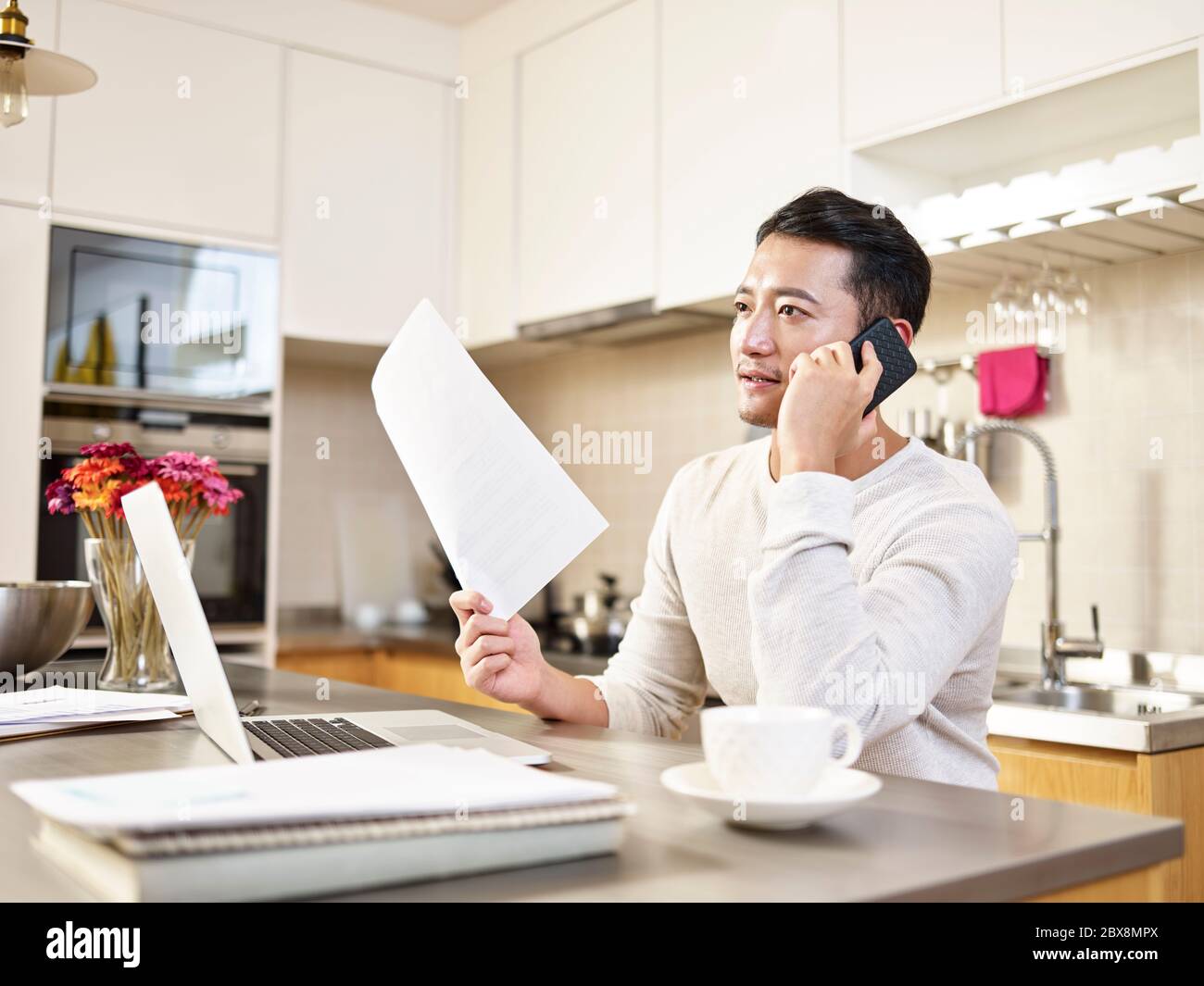 giovane uomo asiatico di affari seduto cucina contatore di lavoro a casa parlando sul telefono cellulare tenere un documento Foto Stock