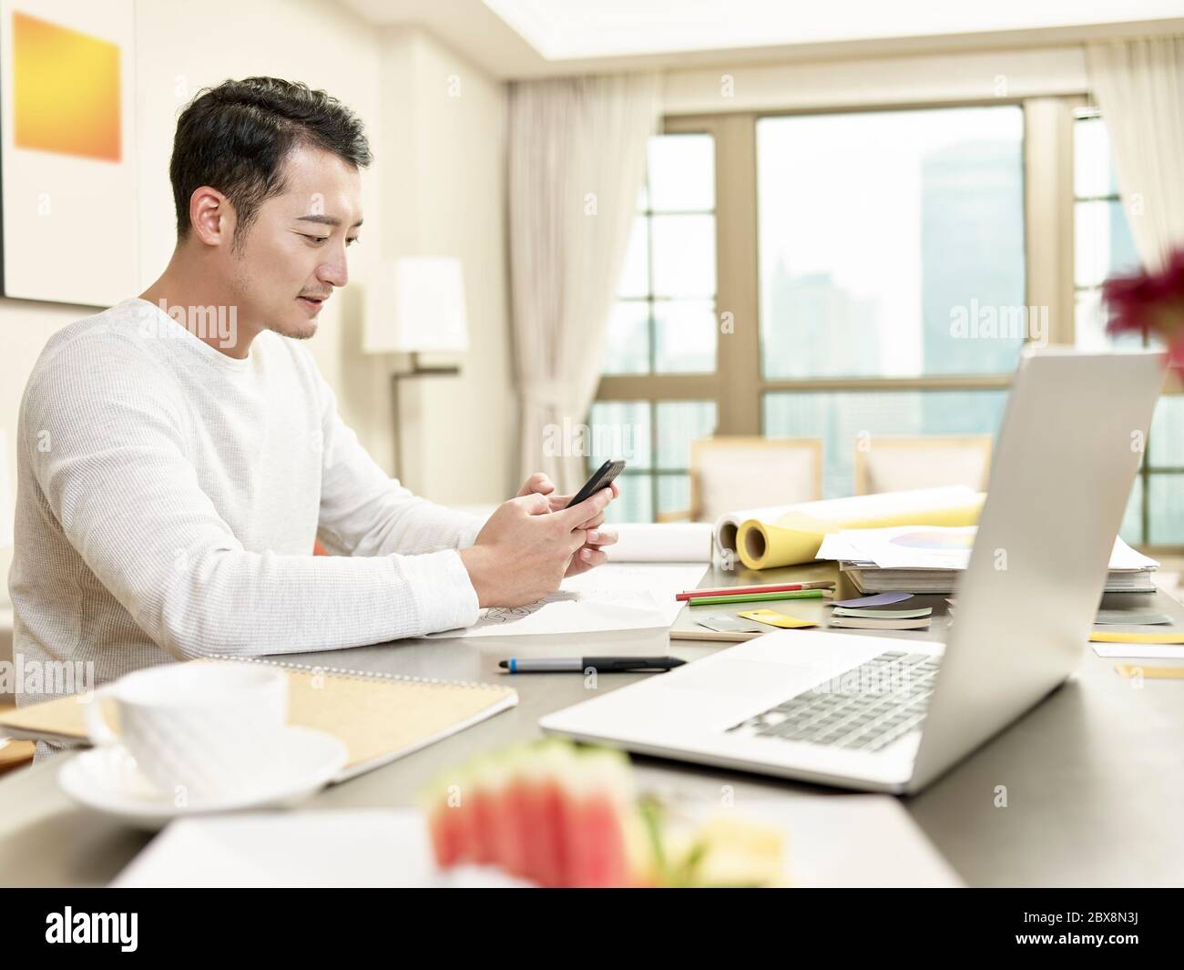 giovane asiatico uomo design professionale di lavoro da casa seduta in cucina utilizzando cellulare (grafica in background alterata digitalmente) Foto Stock