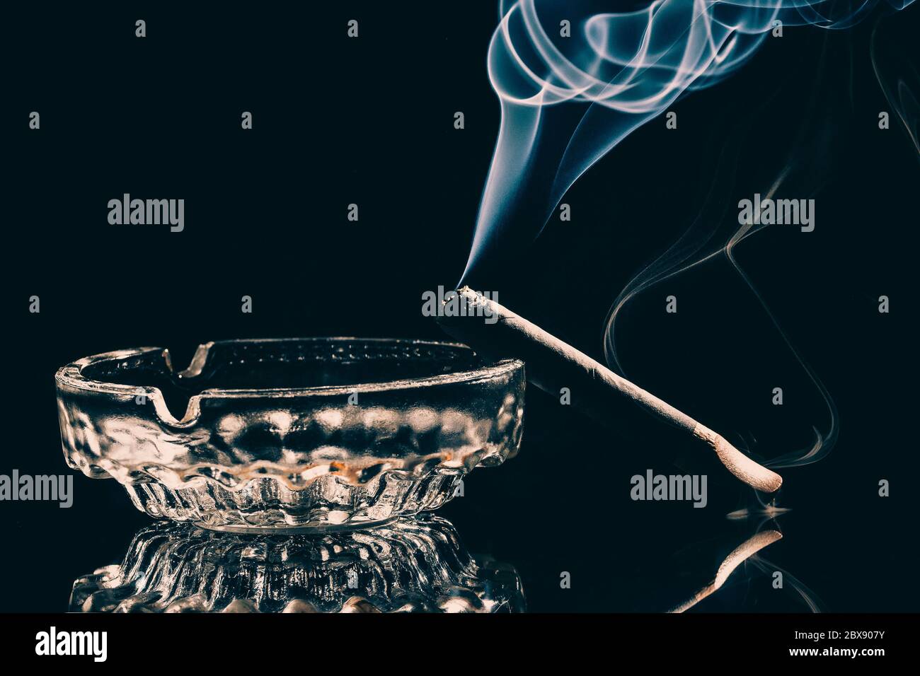 Un sigaro che brucia in un posacenere di vetro con fumo visibile su sfondo nero. Studio girato . Foto tasto basso . Foto Stock