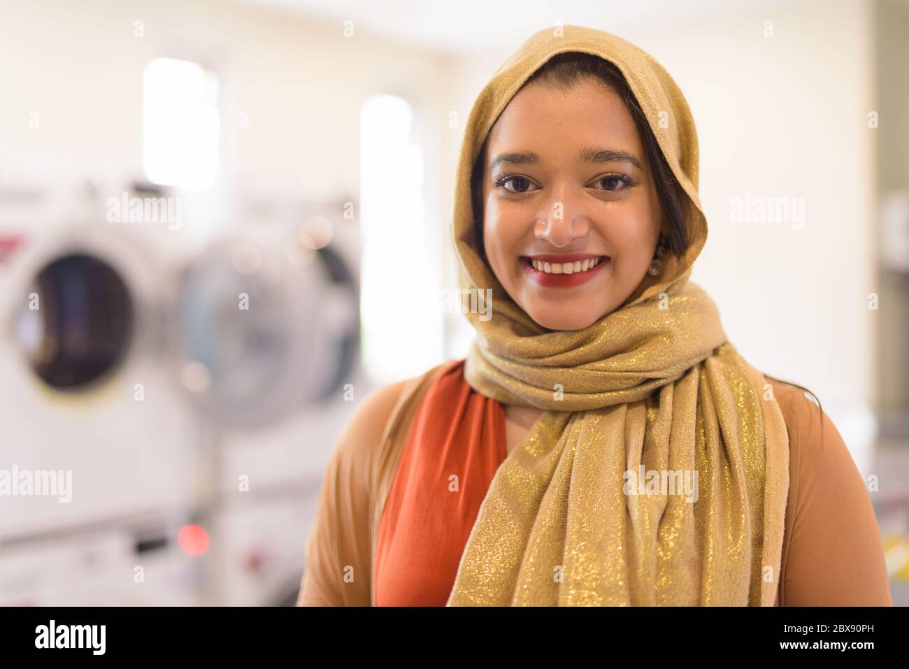 Faccia di felice giovane bella donna d'affari musulmana indiana sorridente come proprietario della lavanderia automatica Foto Stock