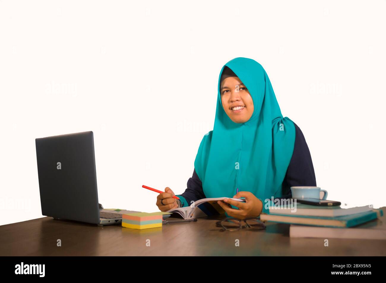Giovane studentessa musulmana felice e di successo nella sciarpa di testa hijab tradizionale Islam che lavora su scrivania studiando con computer portatile e smili libro di testo Foto Stock