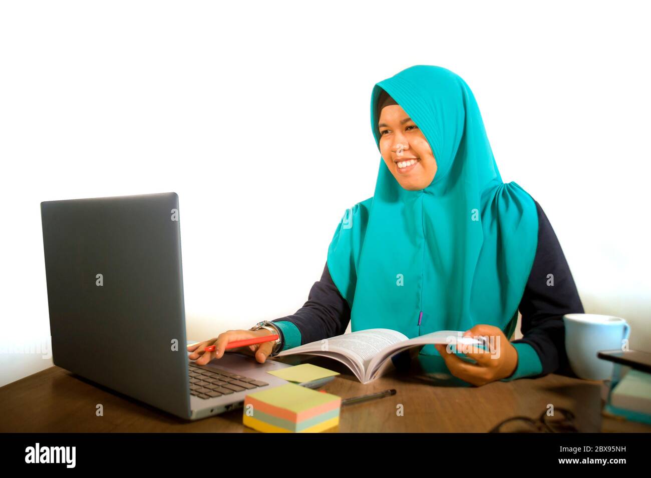 Giovane studentessa musulmana felice e di successo nella sciarpa di testa hijab tradizionale Islam che lavora su scrivania studiando con computer portatile e smili libro di testo Foto Stock