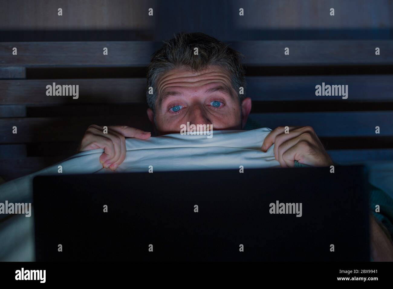 ritratto di stile di vita di giovane paura e stressato uomo a letto guardando film horror internet tarda notte con computer portatile o camera da letto tv in pa Foto Stock