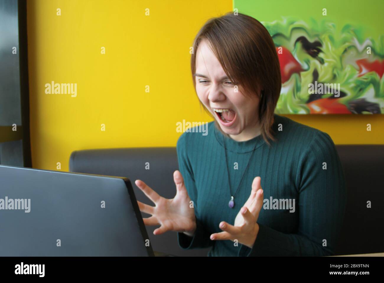 Una ragazza con un'espressione furiosa di tensione sul suo volto si siede lavorando a un computer portatile a un tavolo in un bar su un divano marrone contro una parete gialla. Guarda lo schermo del computer, urla e fa ondate le mani. Foto Stock