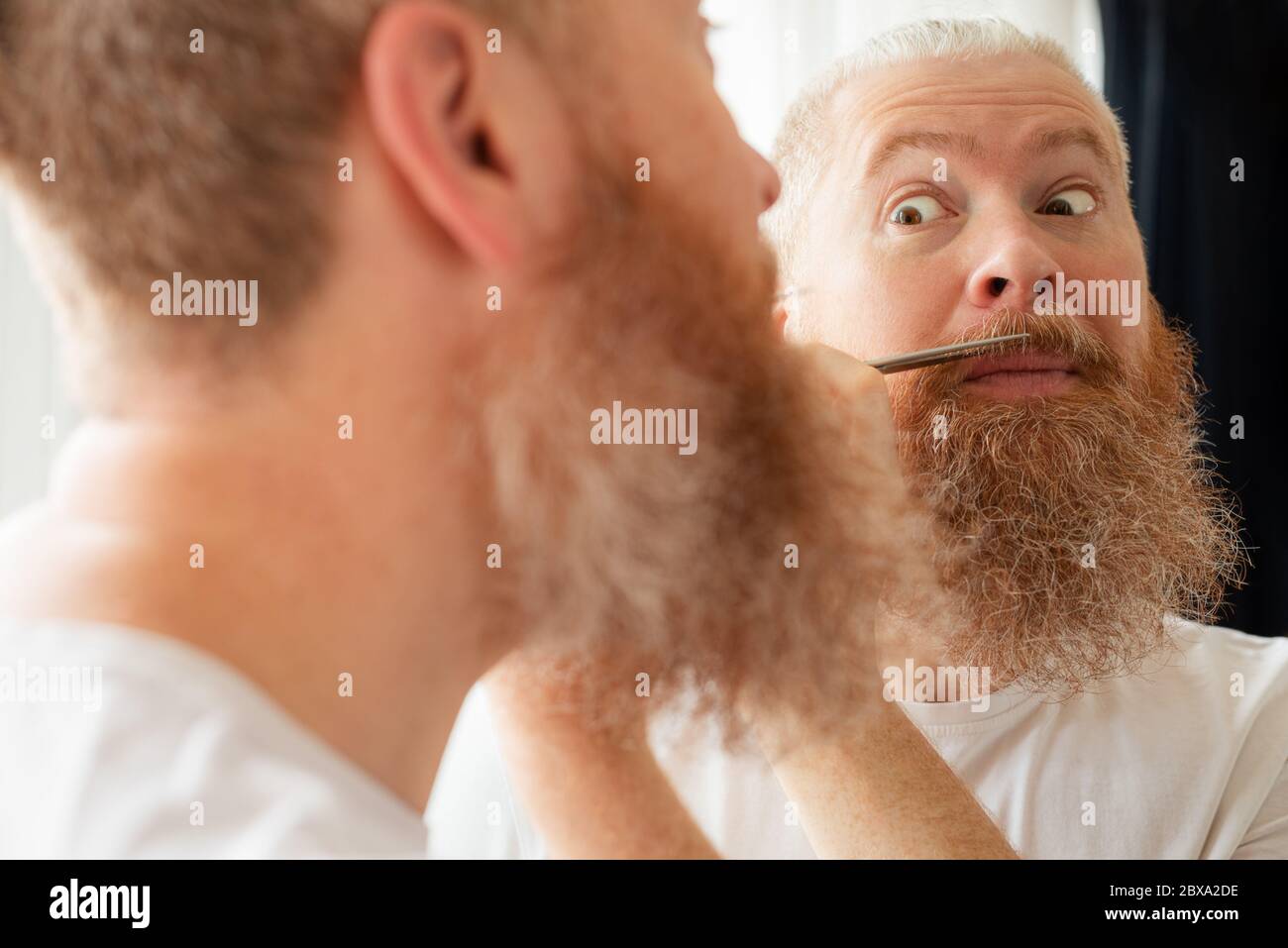 Attraente uomo di mezza età che taglia i baffi e la barba da solo davanti allo specchio a casa. Cura della barba durante la quarantena Foto Stock