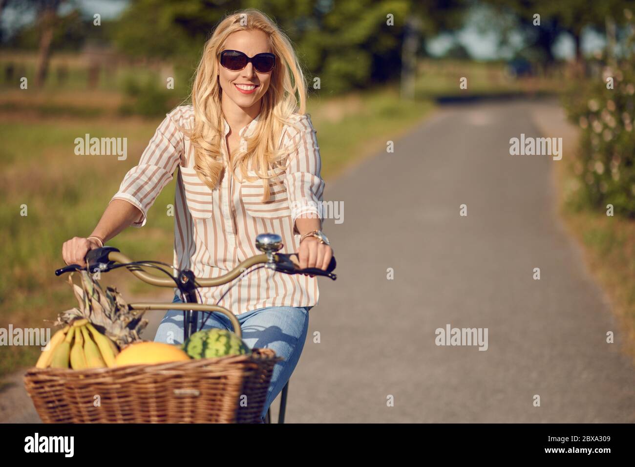 Donna felice sorridente mentre si guida in bicicletta con un cesto pieno di frutta fresca e sana in una giornata di sole d'estate in campagna Foto Stock