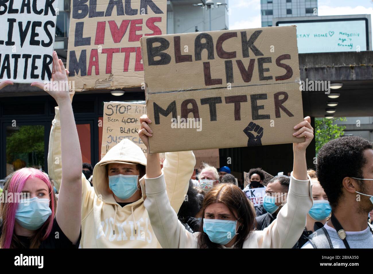 Black Lives Matter protesta Manchester UK. I manifestanti che indossano maschere facciali tengono in alto il cartello. Il messaggio in background sul display visualizza Stay Home Foto Stock