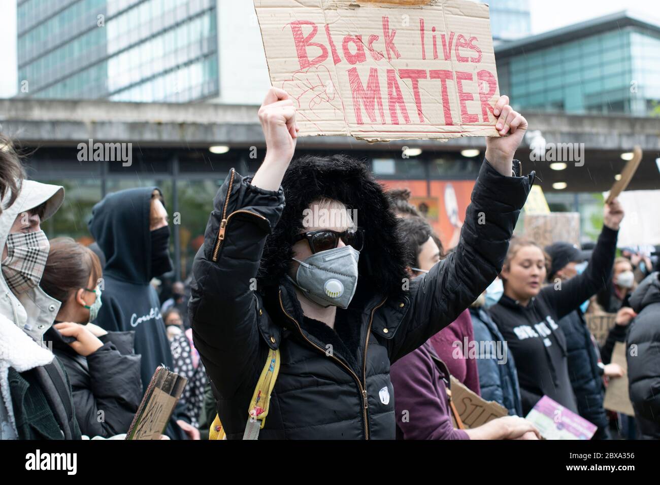 Black Lives Matter protesta Manchester UK. Donna che indossa la maschera tiene in su il segno davanti all'edificio della stazione degli autobus Piccadilly con la folla in background Foto Stock