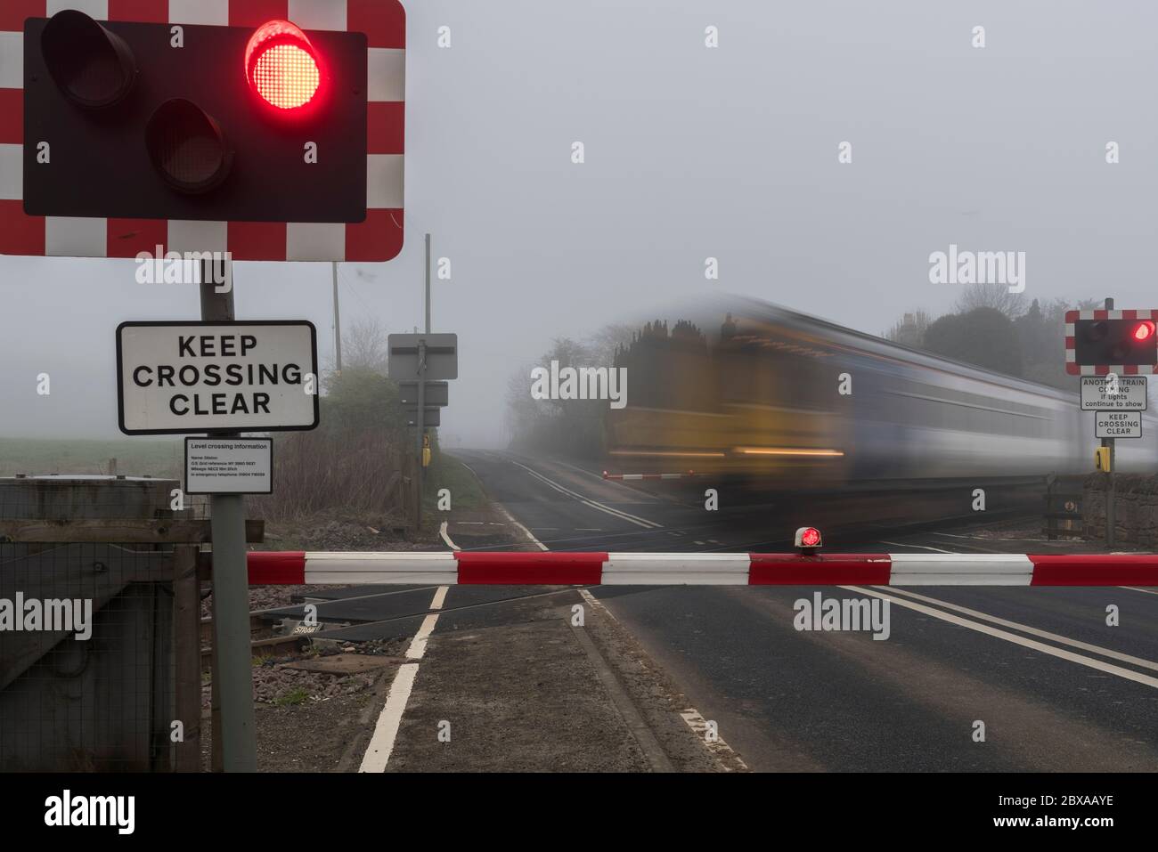 Treno per pendolari di prima mattina che viaggia ad alta velocità attraverso un incrocio a livello stradale con barriere e luci lampeggianti. Foto Stock