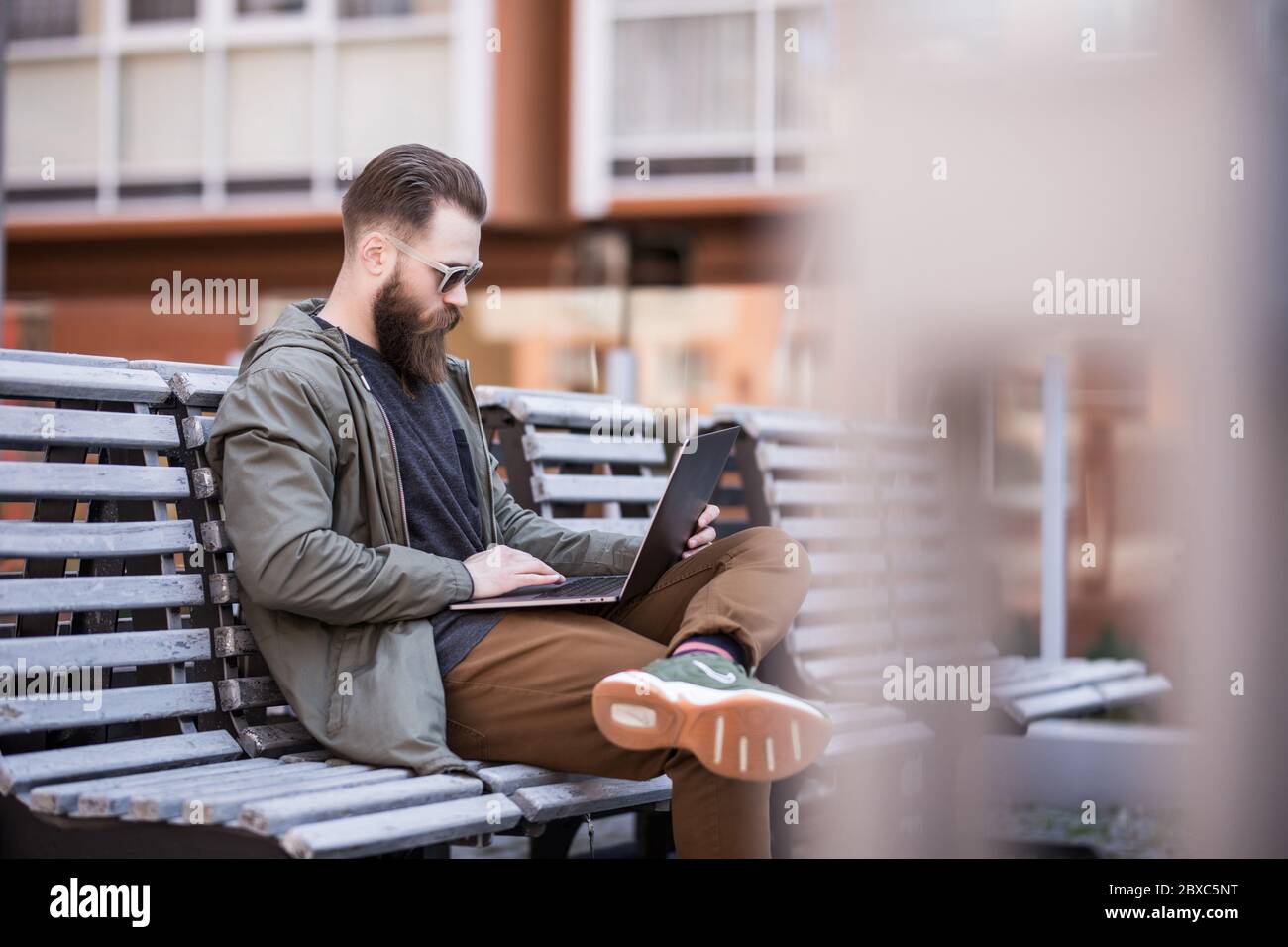 Ritratto di giovane uomo bearded alla strada. Stile di vita con un bel ragazzo all'aperto sulla panca che lavora con un moderno notebook grigio all'aperto Foto Stock