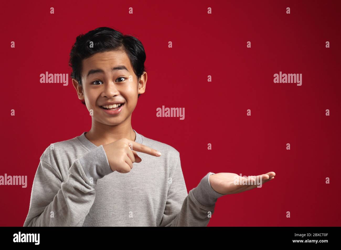 Cute bambino asiatico ragazzo con palma aperta e dito puntato in su. Promuovere la presentazione di qualcosa, copiare spazio su sfondo rosso Foto Stock