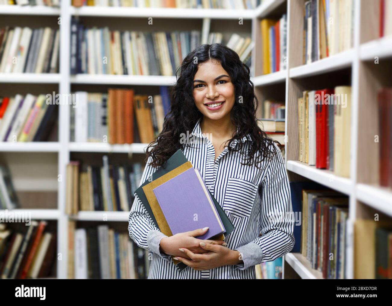 Ragazza latina sorridente che tiene libri di testo alla biblioteca Foto Stock