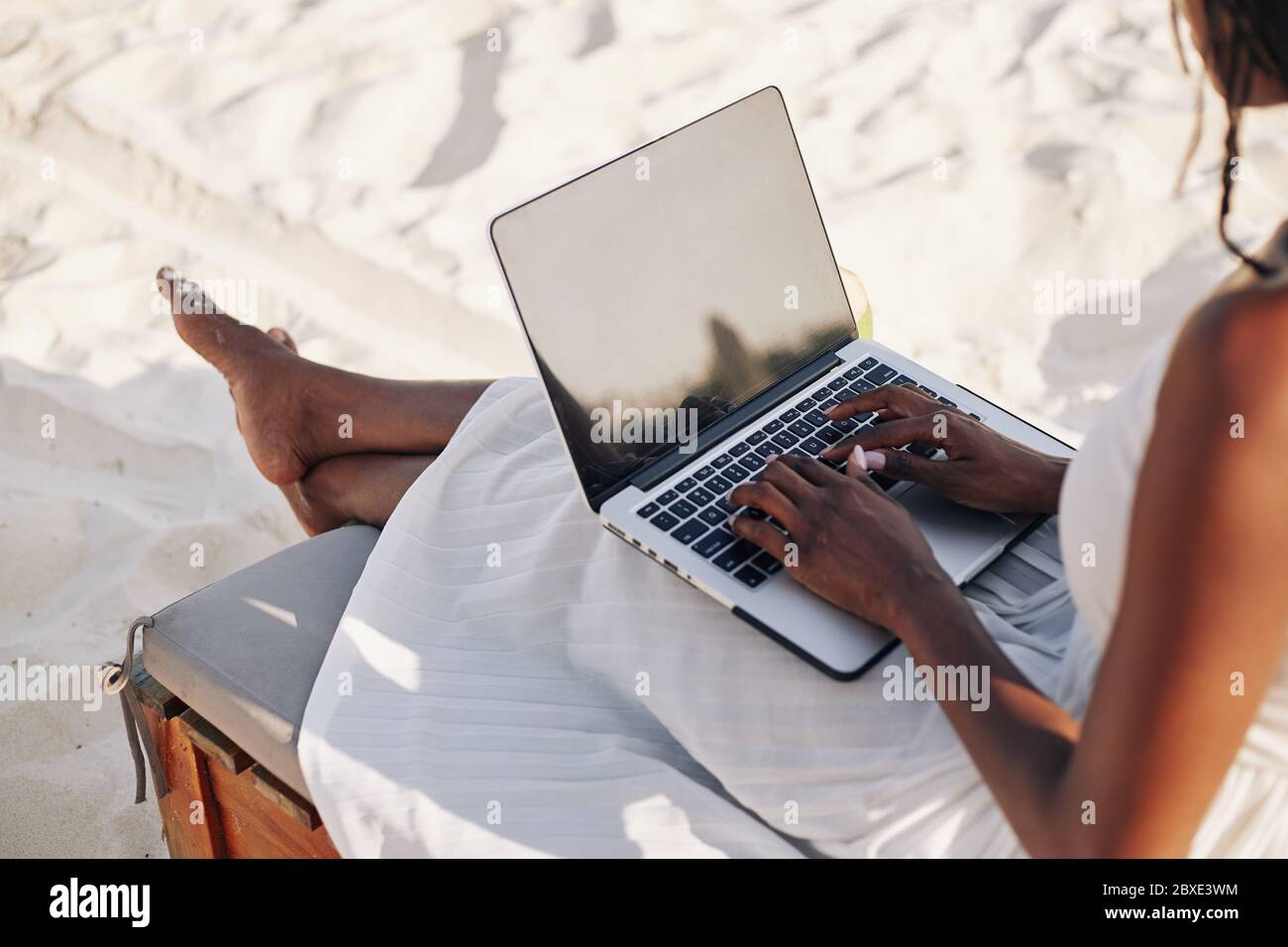 Giovane donna con computer portatile seduto sulla chaise-lounge sulla spiaggia e lavorando a distanza Foto Stock