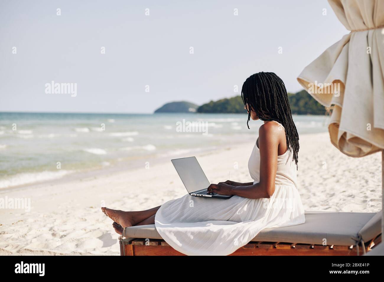 Bella giovane donna con dreadlock seduta sulla chaise-lounge, lavorando su un computer portatile e godendo il suono delle onde Foto Stock