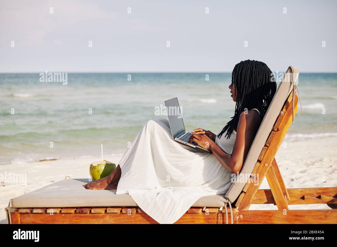 Giovane donna che si gode seduta sulla chaise-lounge, lavorando su un computer portatile e guardando le onde del mare Foto Stock