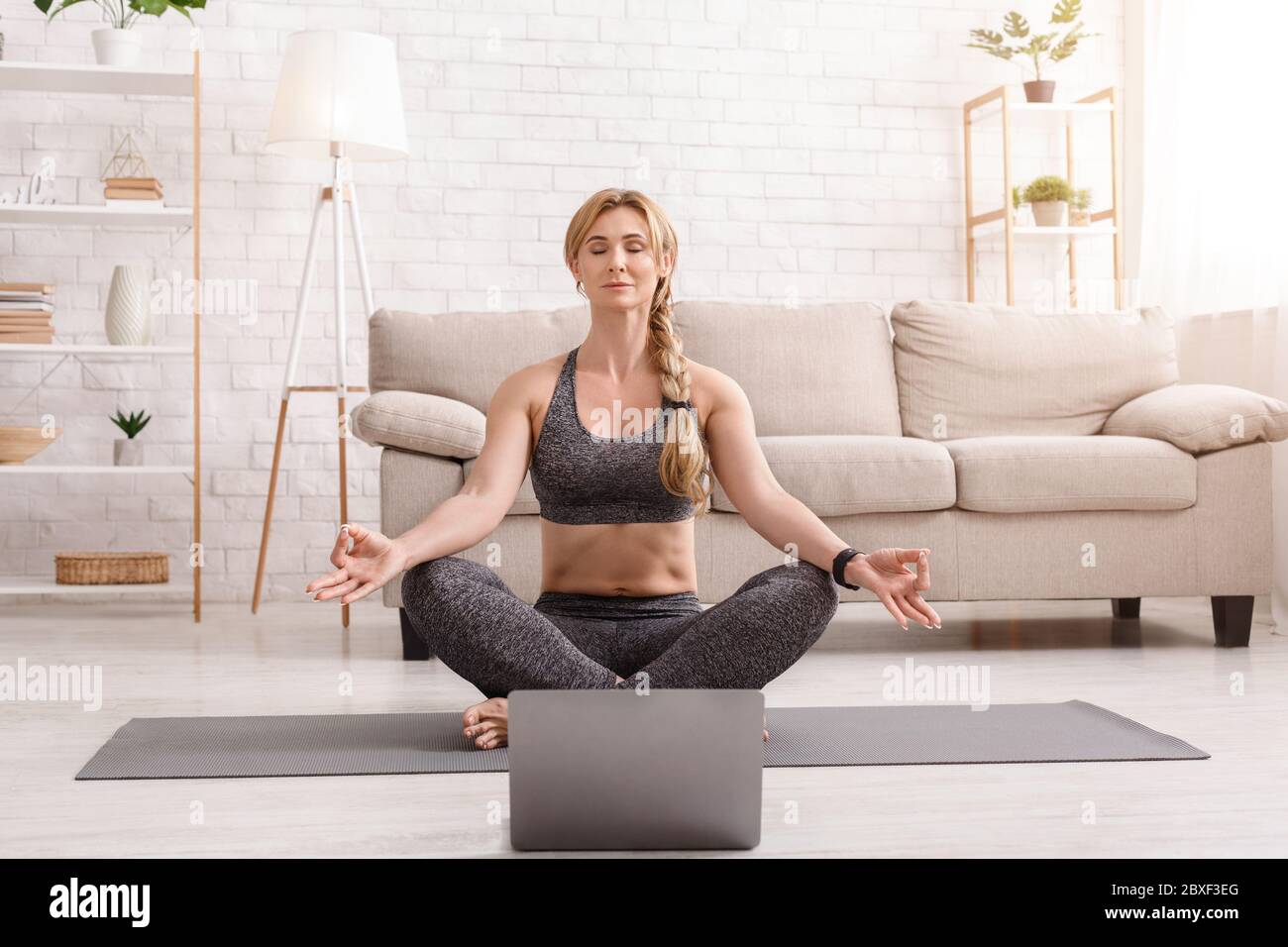 Yoga e meditazione a casa. Donna in posa lotus, seduta sul tappetino davanti al laptop Foto Stock